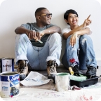 Couple sits on floor surrounded by paint buckets, woman smiles and points while explaining something. 